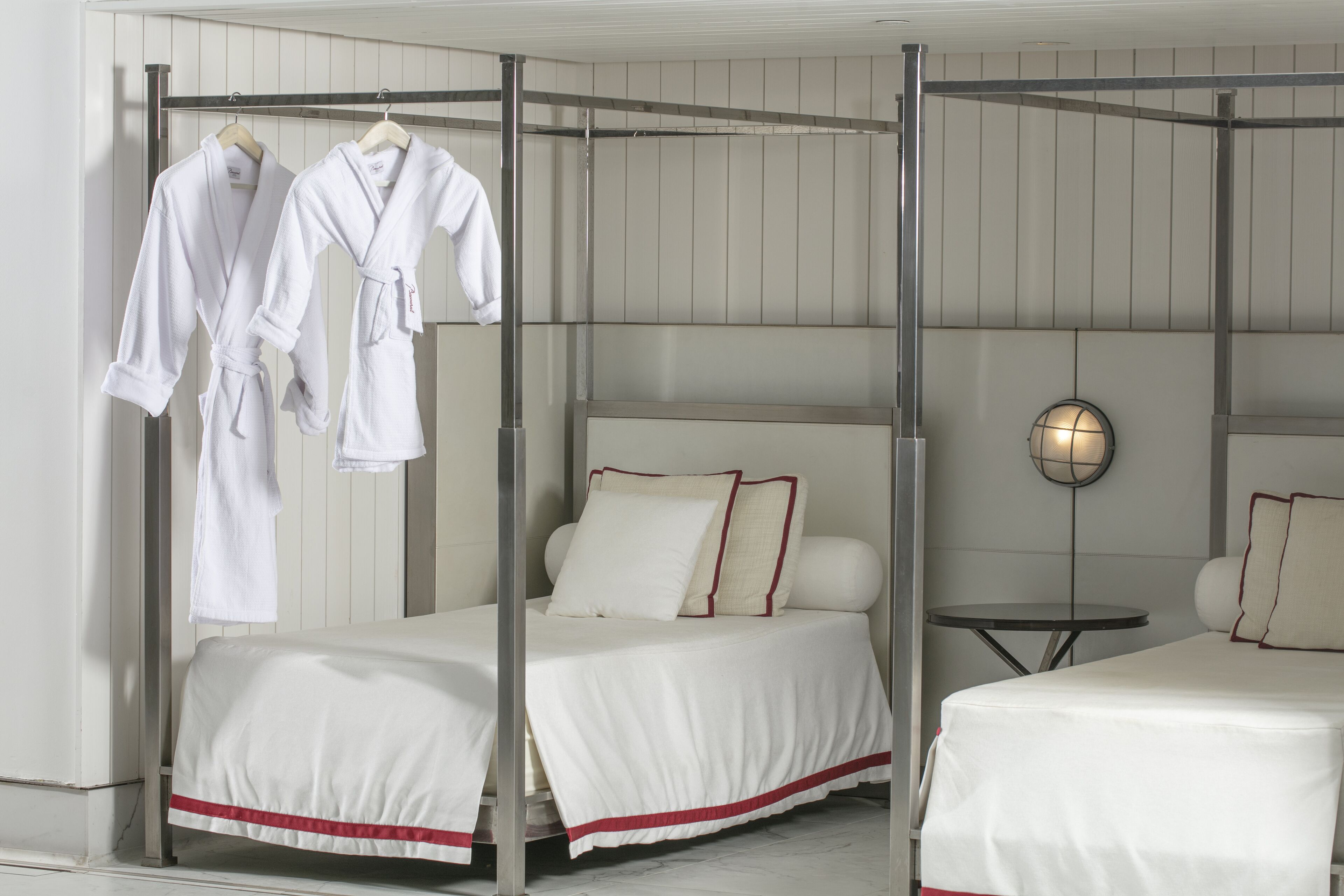A hotel room with two beds, each with white bedding and red trim, a metal canopy bed, a round wall-mounted lamp, and a small round table.