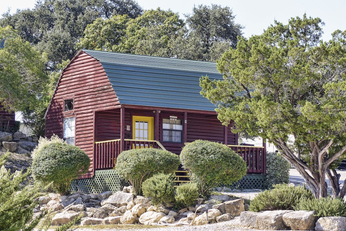 A rustic log cabin with a green roof, a yellow front door, and a porch with a railing.