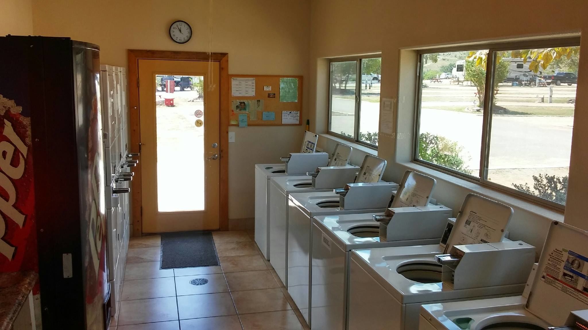 A laundry room with multiple washing machines, a clock on the wall, and a bulletin board with notices.