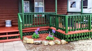 Una cabaña de madera con un porche verde, un pequeño jardín con rocas y flores púrpuras, y un techo de metal.
