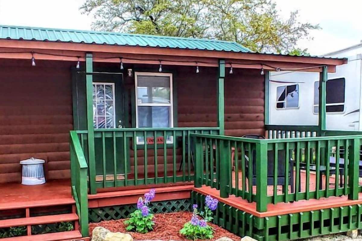 A wooden cabin with a green porch, a small garden with rocks and purple flowers, and a metal roof.