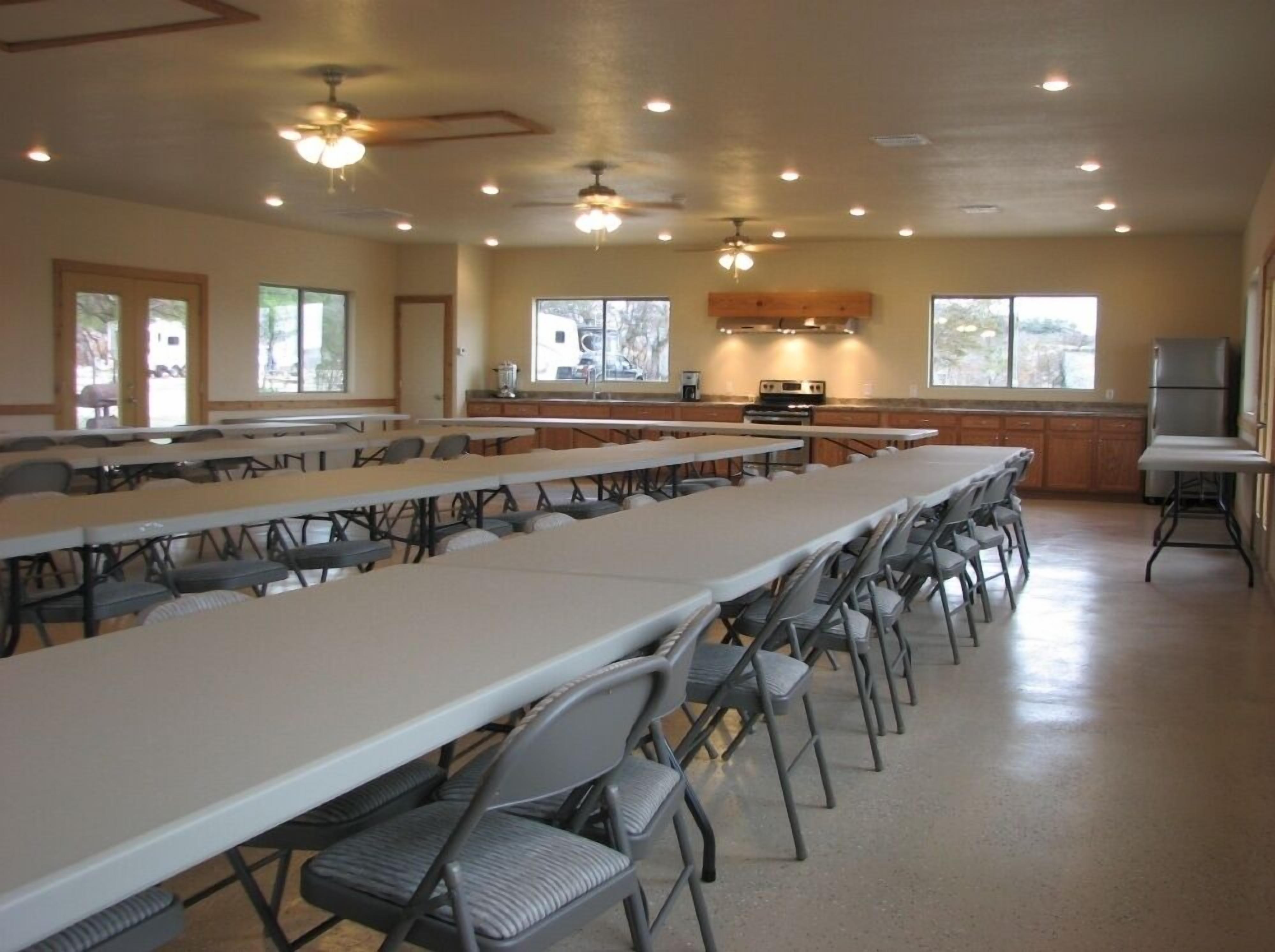 A large conference room with long tables and chairs, a kitchen area with a refrigerator, and a ceiling fan.