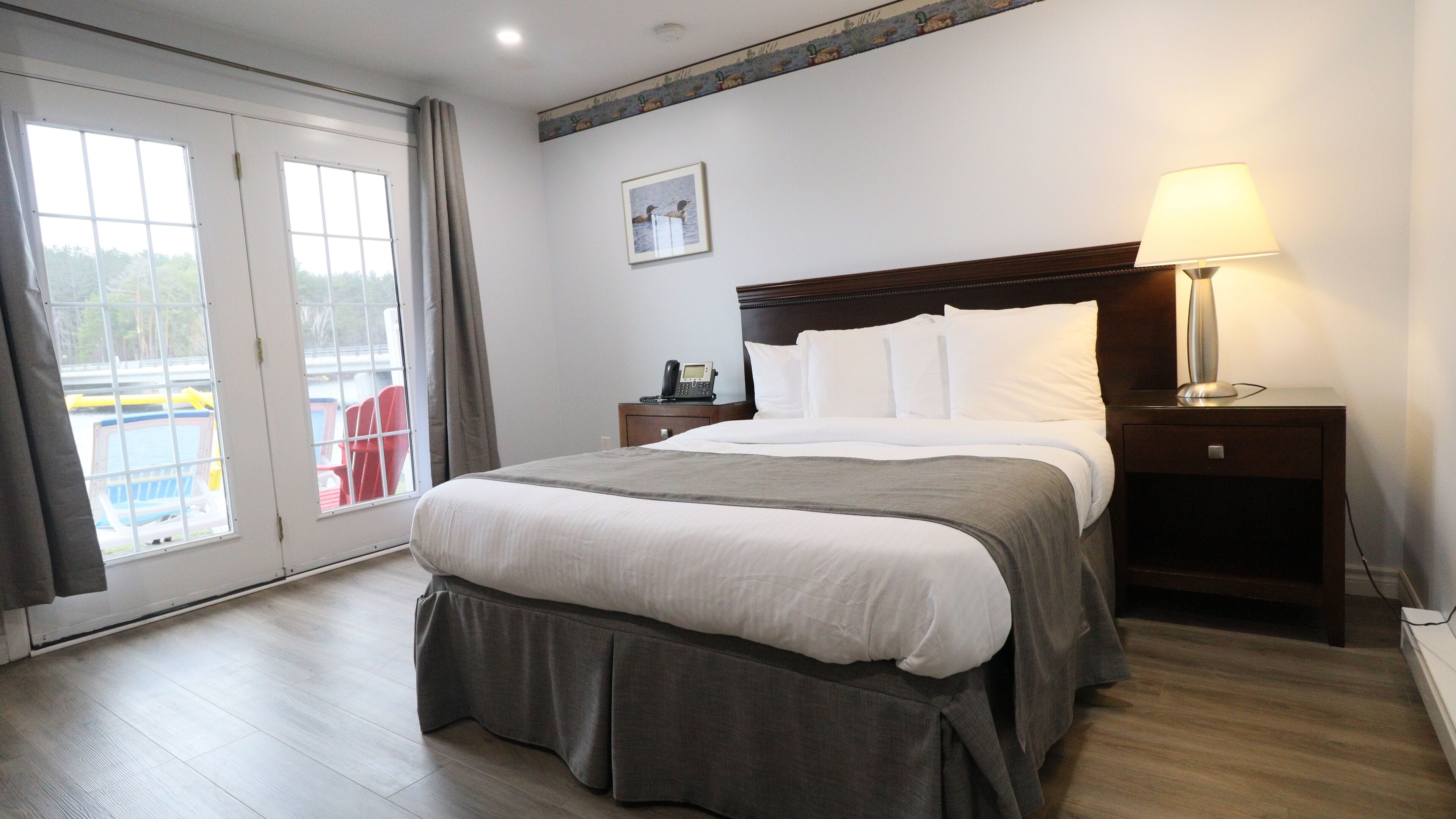 Une chambre d’hôtel avec un grand lit, une table de chevet avec une lampe, un téléphone et une vue sur une piscine et des chaises, visible à travers une porte-fenêtre coulissante.