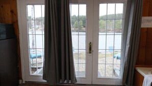 A room with a wooden interior, a door with a view of a lake, and a small outdoor seating area with chairs.