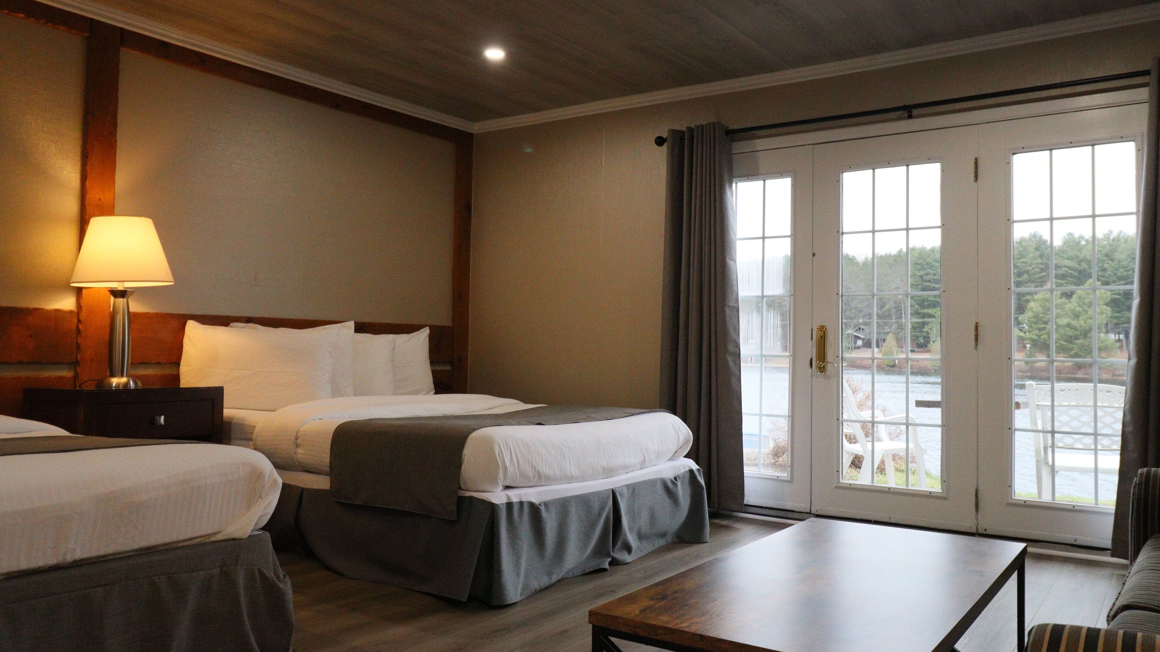 A hotel room with two beds, a desk, a chair, and a view of a lake through the glass doors.