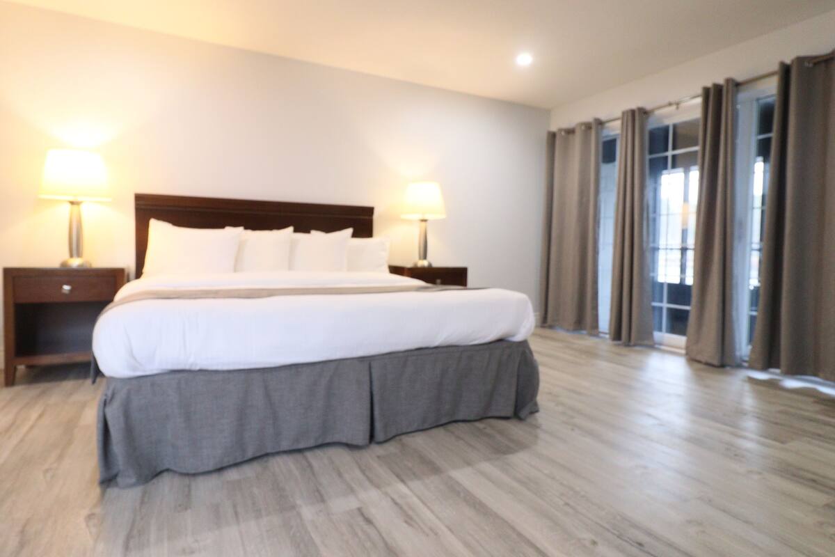 Une chambre d’hôtel avec un grand lit, deux tables de chevet avec des lampes, une tête de lit en bois, un lit recouvert d’une housse grise et une vue sur l’extérieur par une fenêtre avec des rideaux.