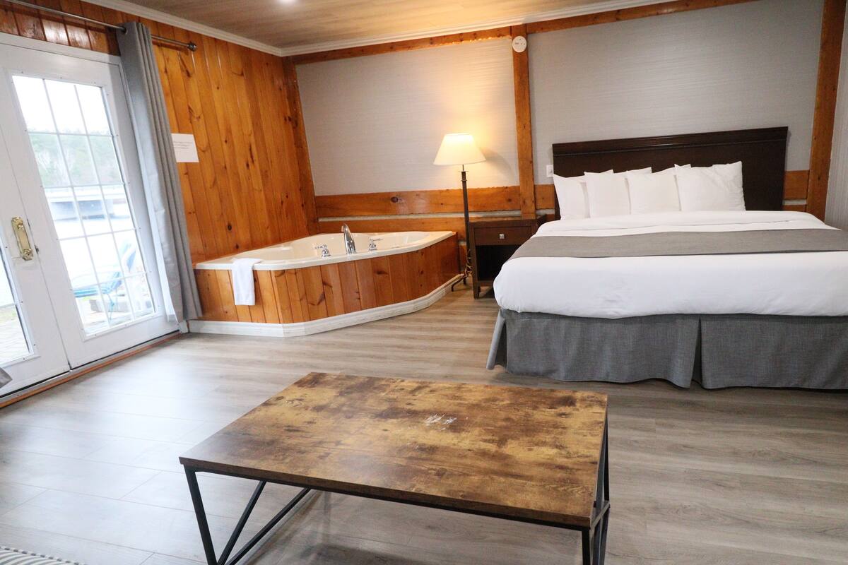 Une chambre d’hôtel avec un plancher en bois, un lit avec une literie blanche, une petite table, une baignoire et une lampe.