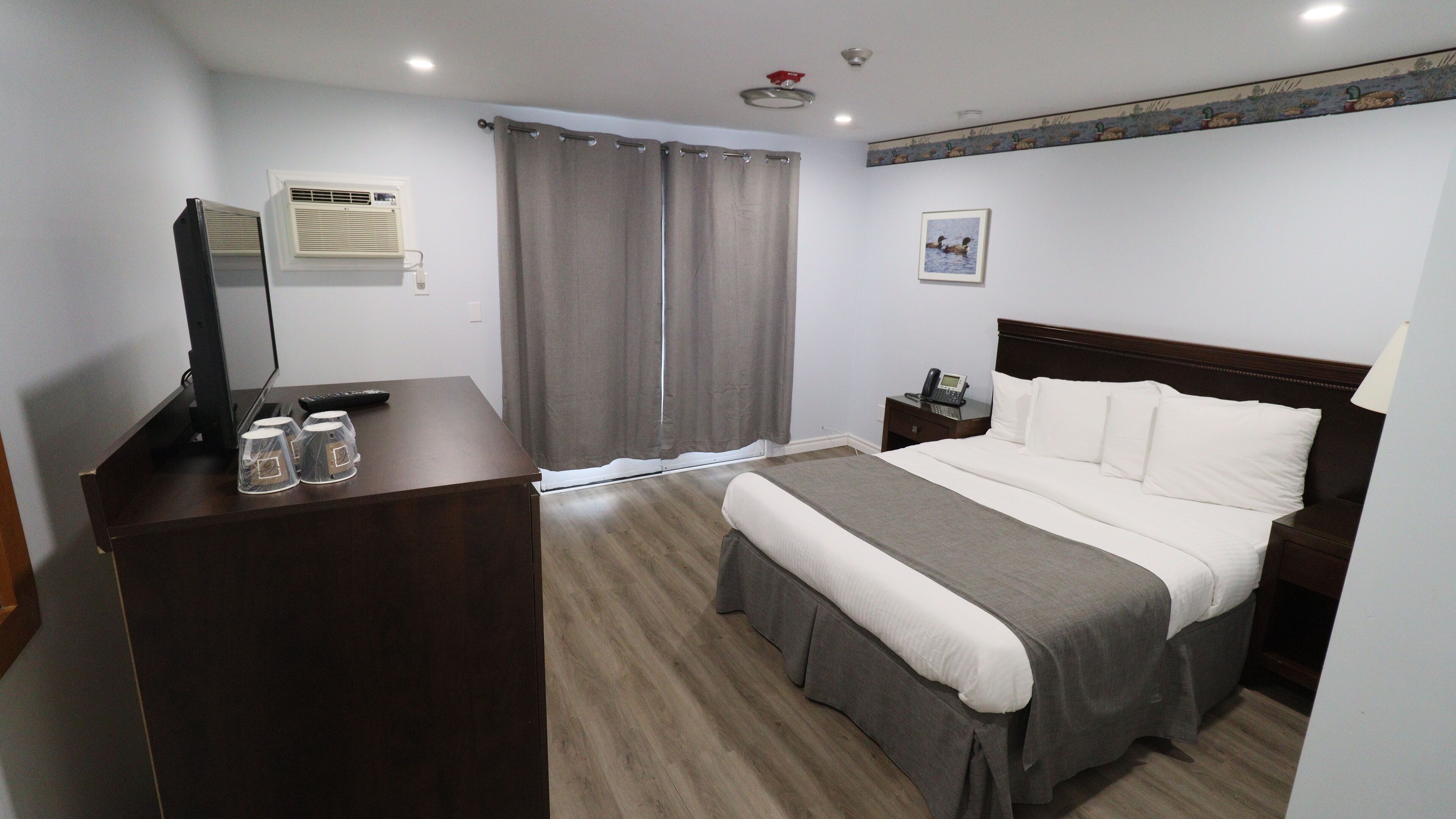 A hotel room with a bed, bedside table, lamp, desk with a computer, and a window with curtains.