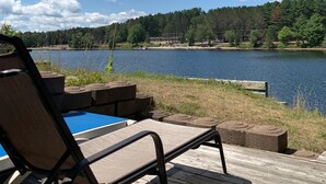 A wooden deck with a lounge chair and a small pool, overlooking a lake with a forested shoreline.