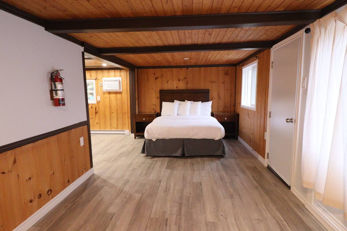Une chambre avec un plafond en bois, un lit avec une literie blanche, une table de chevet, un extincteur fixé au mur et une fenêtre avec des rideaux.