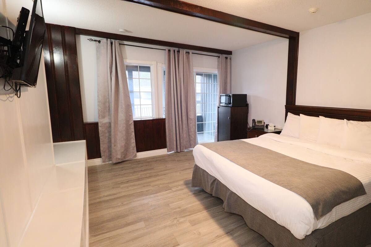 Une chambre d’hôtel avec un grand lit, un plancher en bois, un téléviseur fixé au mur et une fenêtre avec des rideaux.