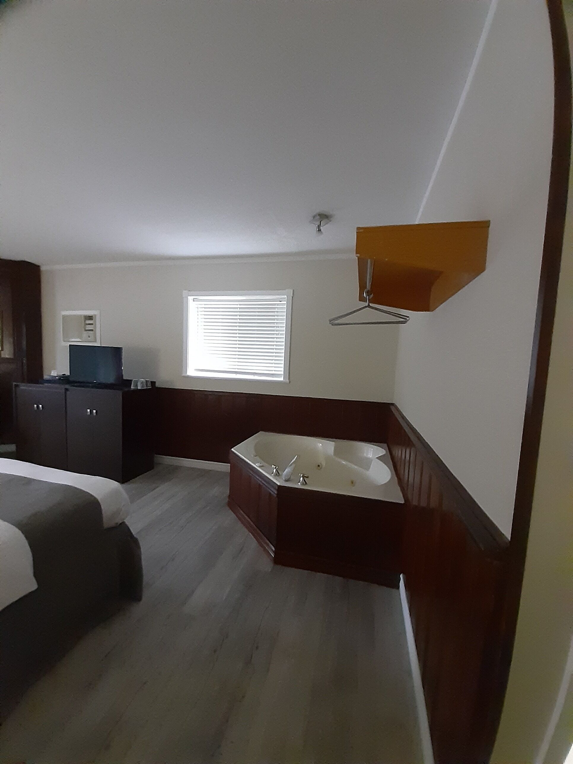 A hotel room with a bed, a desk, a chair, a window with blinds, a hanging light fixture, and a bathtub.