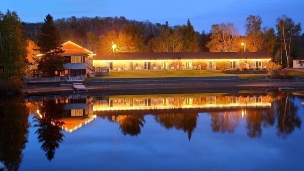 A lodge with a large building and a smaller structure, both illuminated, reflecting on a calm body of water, surrounded by trees.