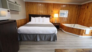 A hotel room with a large bed, a wooden panel wall, a built-in bathtub, and a bedside table with a lamp.