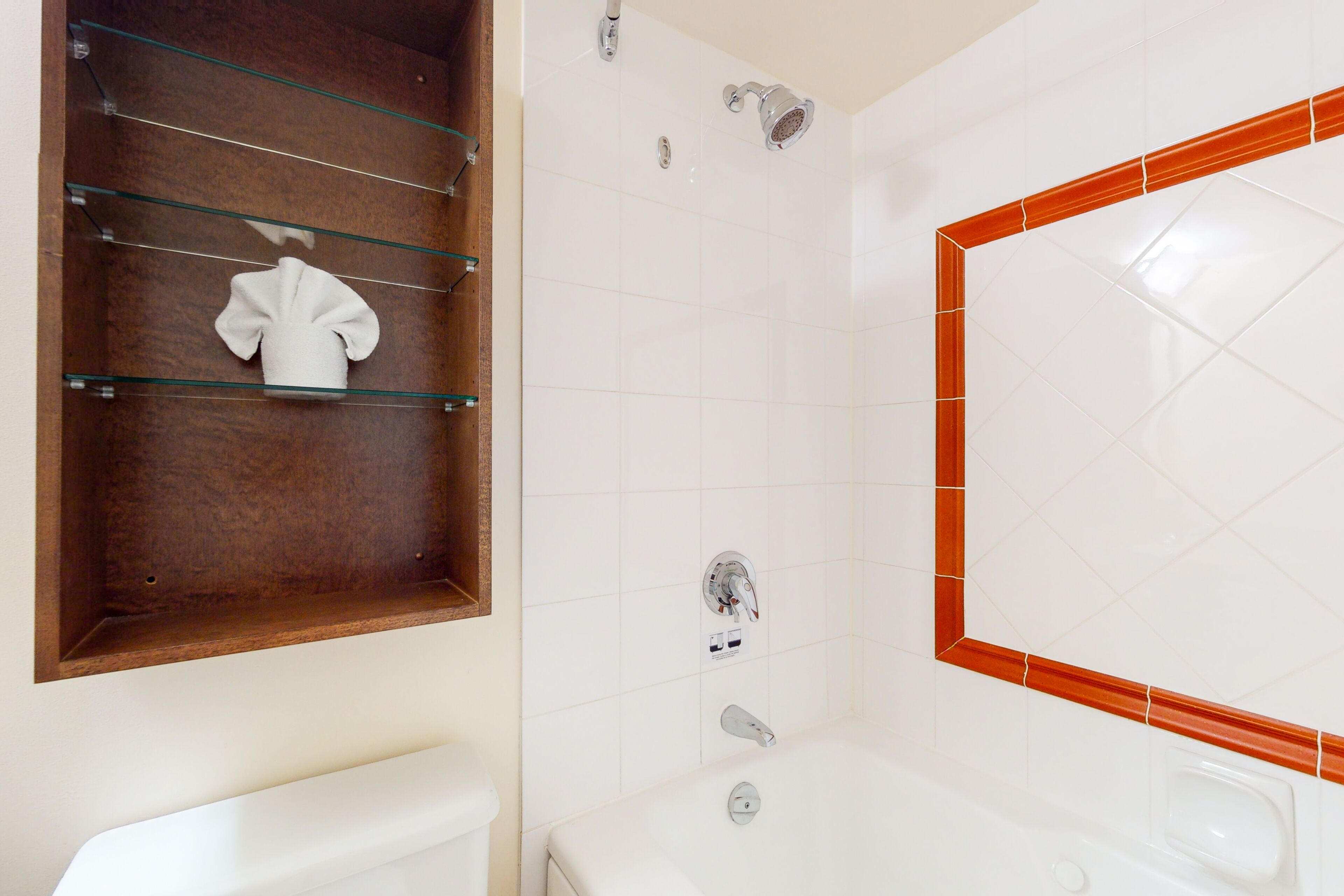 A bathroom with a toilet, a glass shelf with a towel, a shower with a wall-mounted faucet, and a bathtub with a tiled surround.