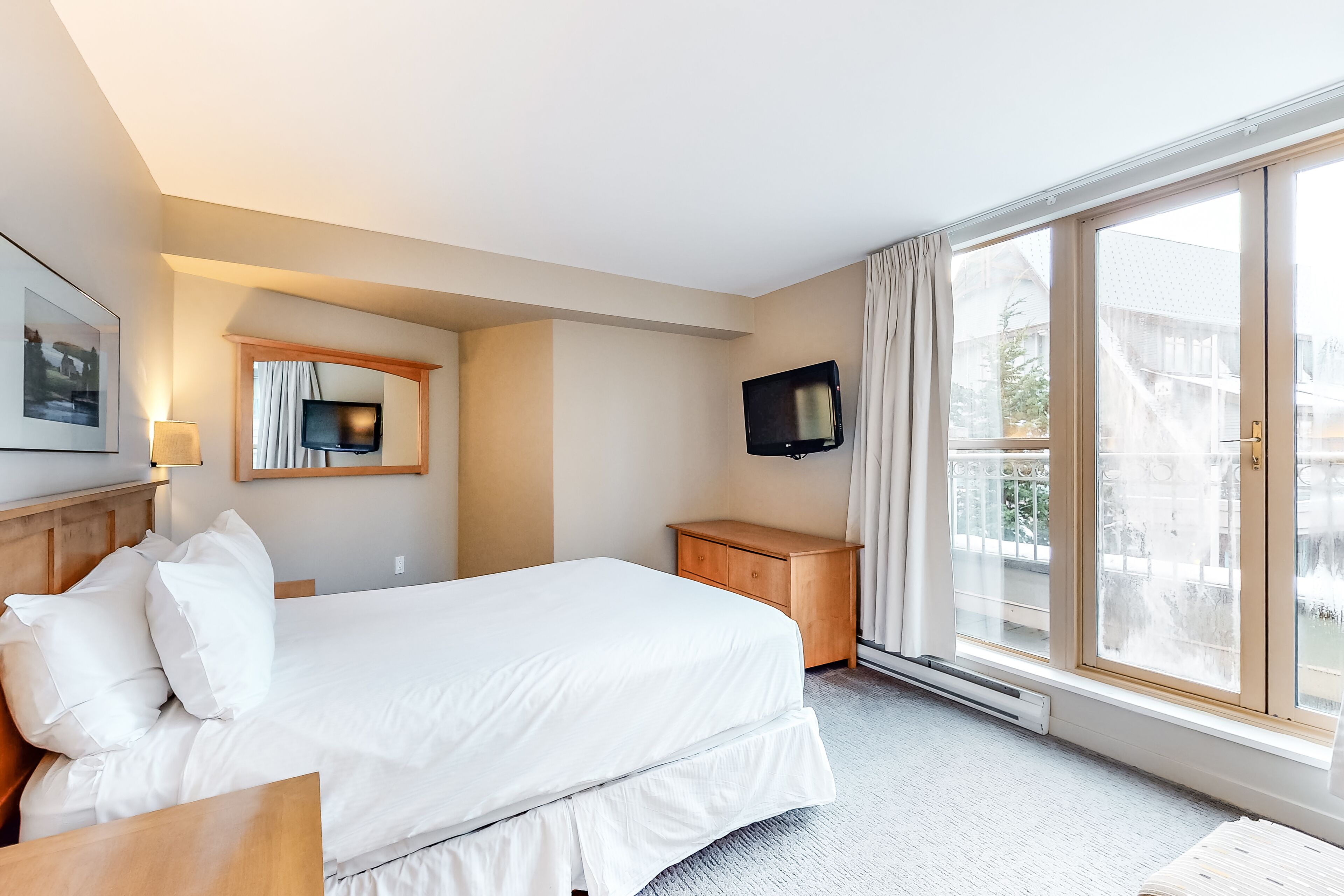 A hotel room with a large bed, a wooden nightstand, a TV mounted on the wall, and a large window with curtains.