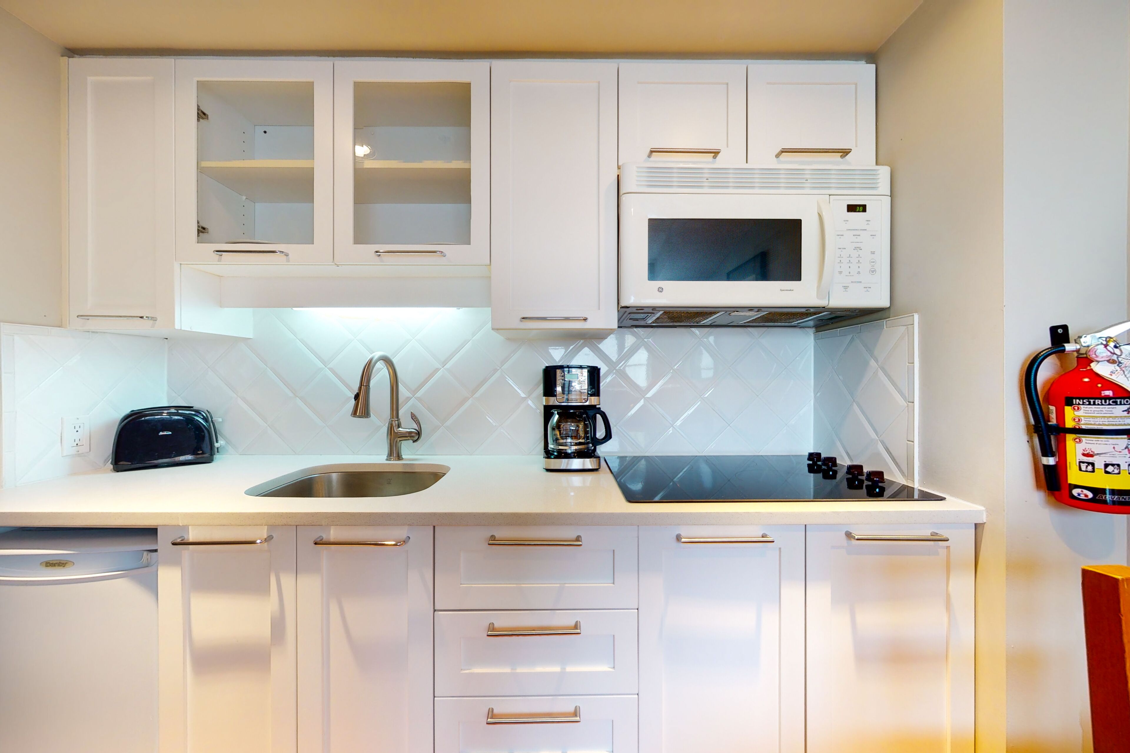 Una cucina con mobili bianchi, un forno a microonde, un tostapane, un lavello e un estintore.