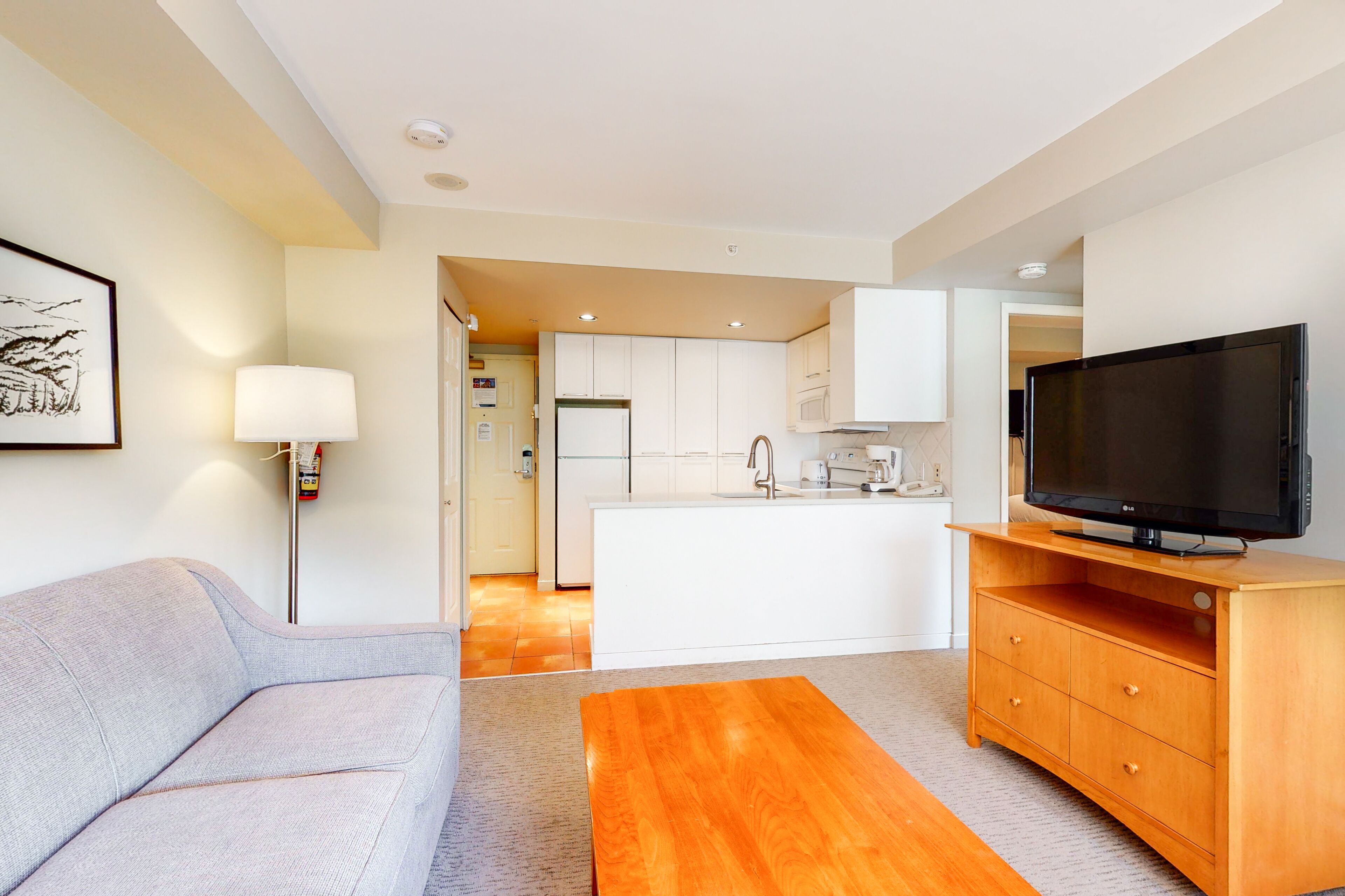 A modern living room with a sofa, a wooden coffee table, a TV on a wooden stand, and a kitchen in the background.