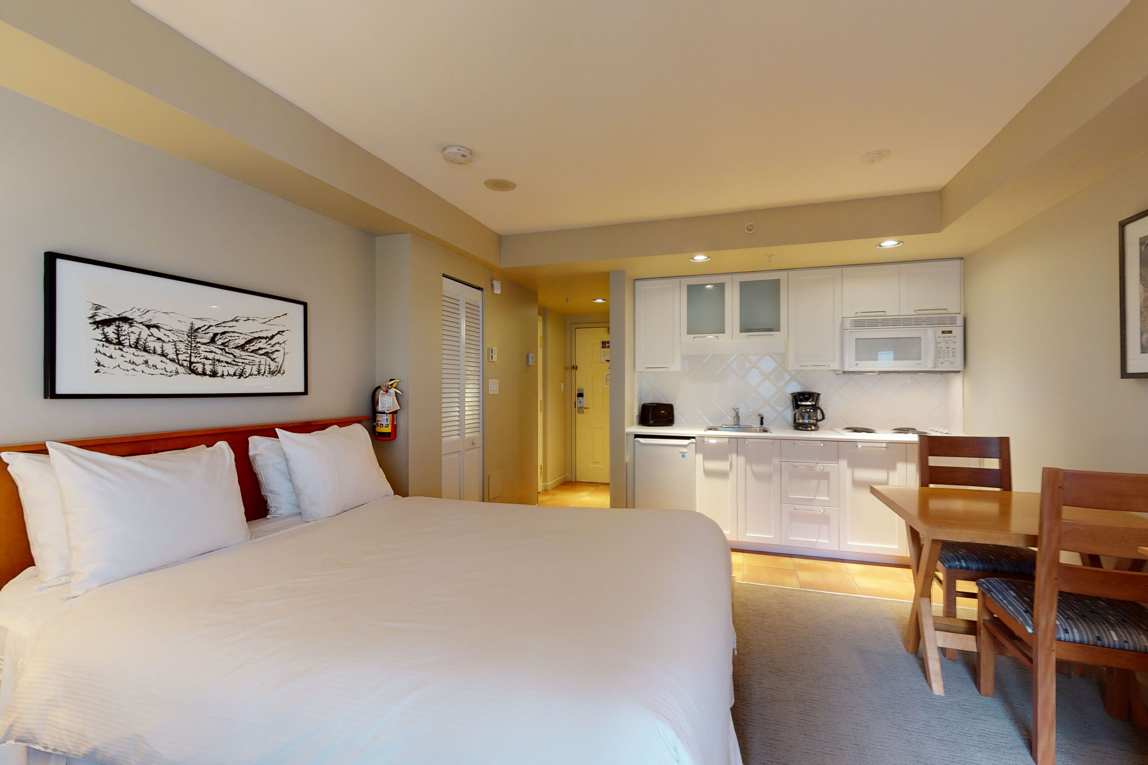 A hotel room with a large bed, a wooden chair, a small table, a kitchenette with a microwave, and a framed picture on the wall.