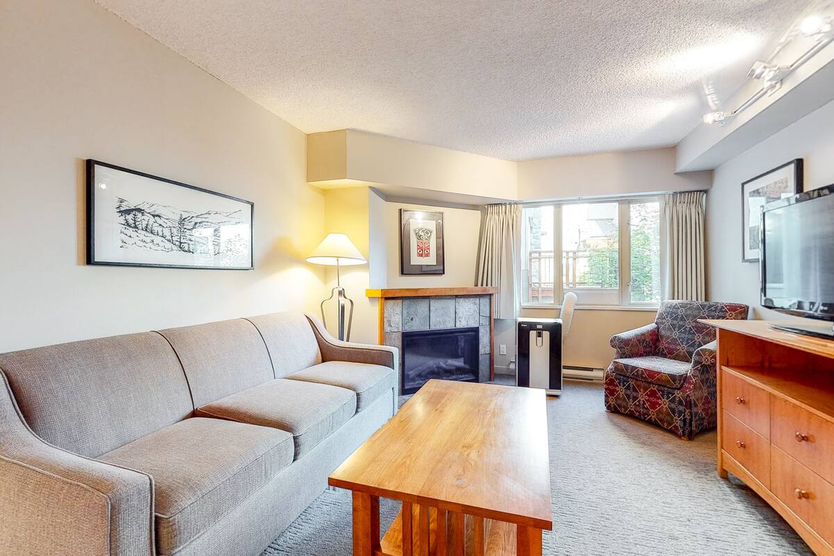 A living room with a sofa, a wooden coffee table, a patterned armchair, a fireplace, a television, and a large window with curtains.