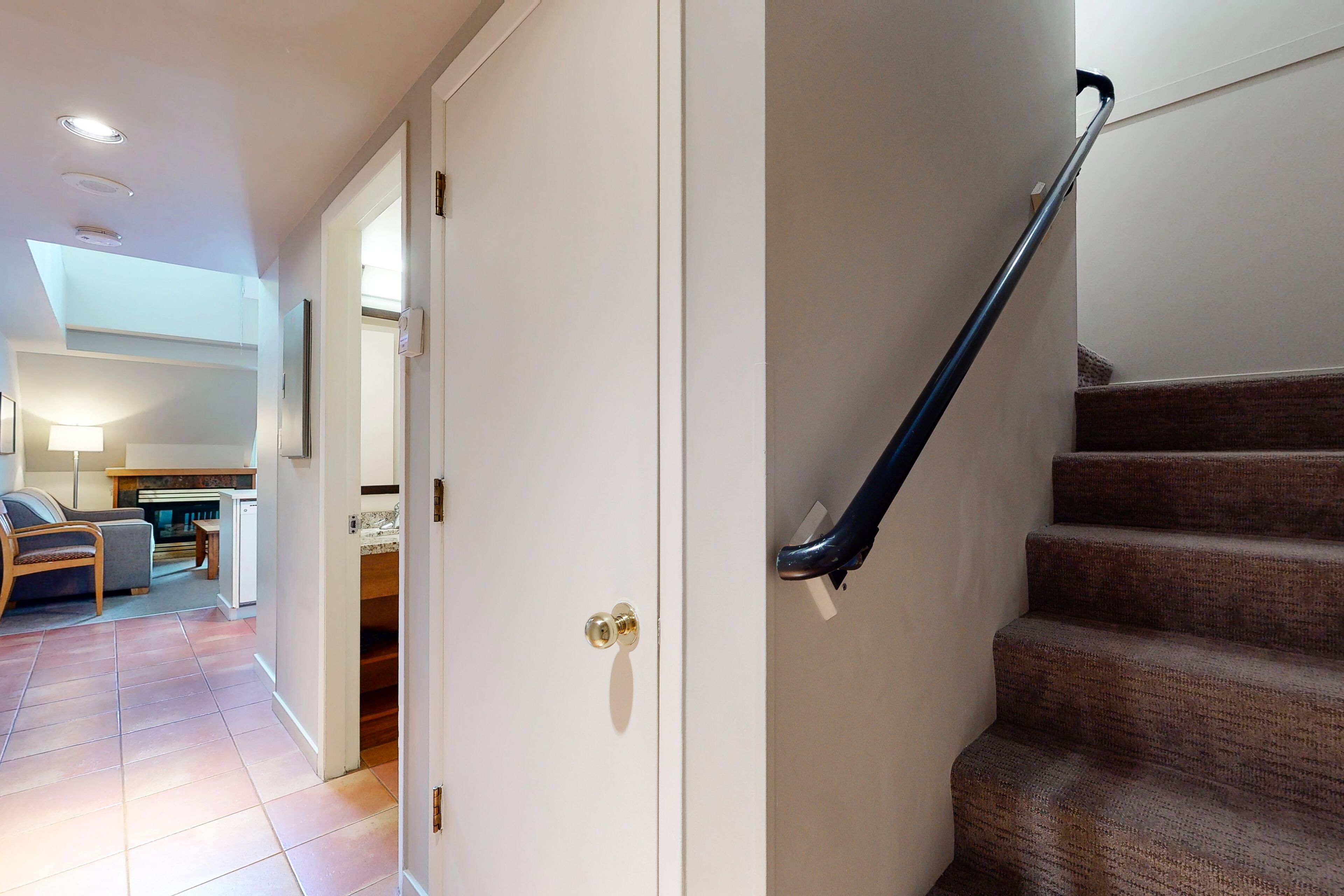 A staircase with a handrail, a door with a brass handle, and a view into a living area with a sofa and a lamp.