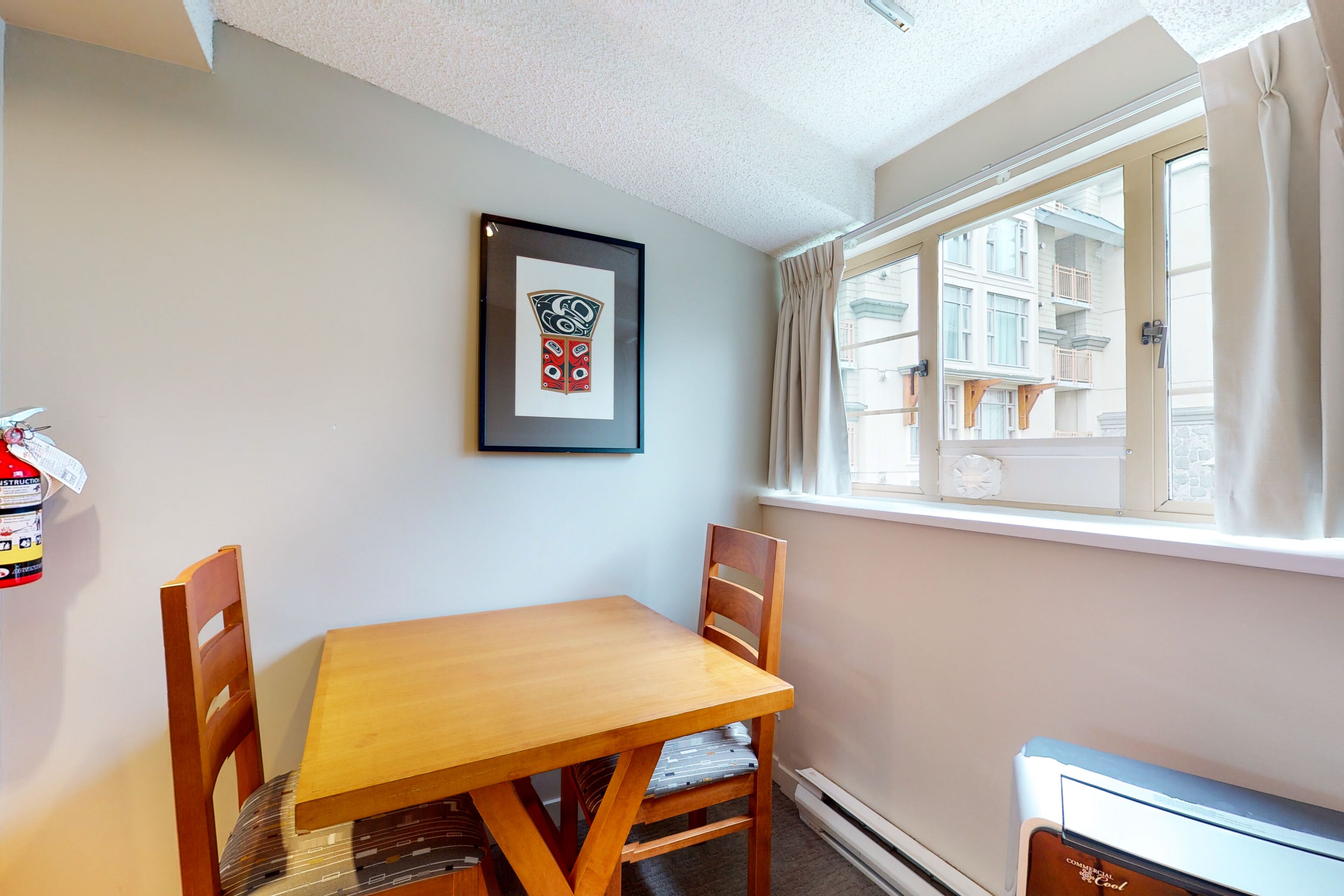 Una habitación con una mesa y sillas de madera, un cuadro en la pared, una ventana con cortinas y un radiador.