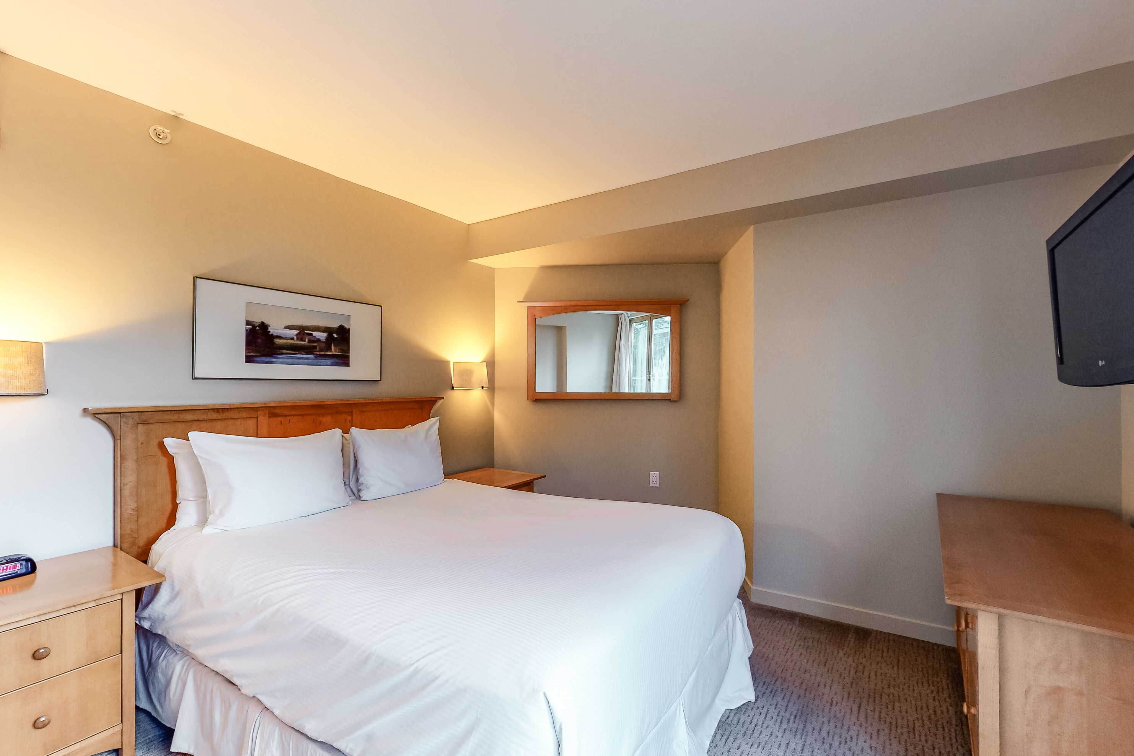 A hotel room with a large bed, a wooden headboard, a bedside table with a lamp, a framed picture on the wall, and a mirror.