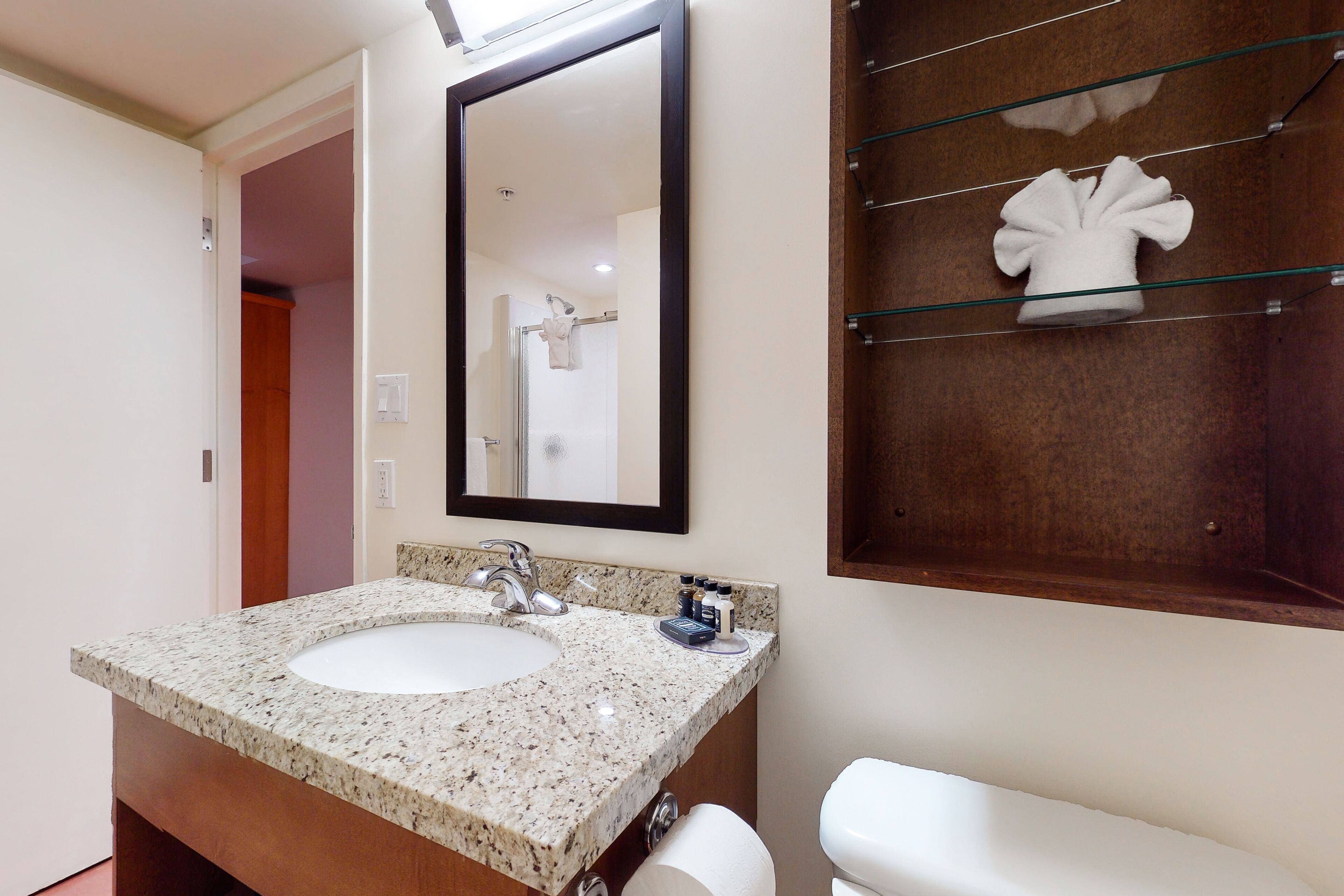 A bathroom with a granite countertop sink, a mirror, and a glass shelf with a towel.