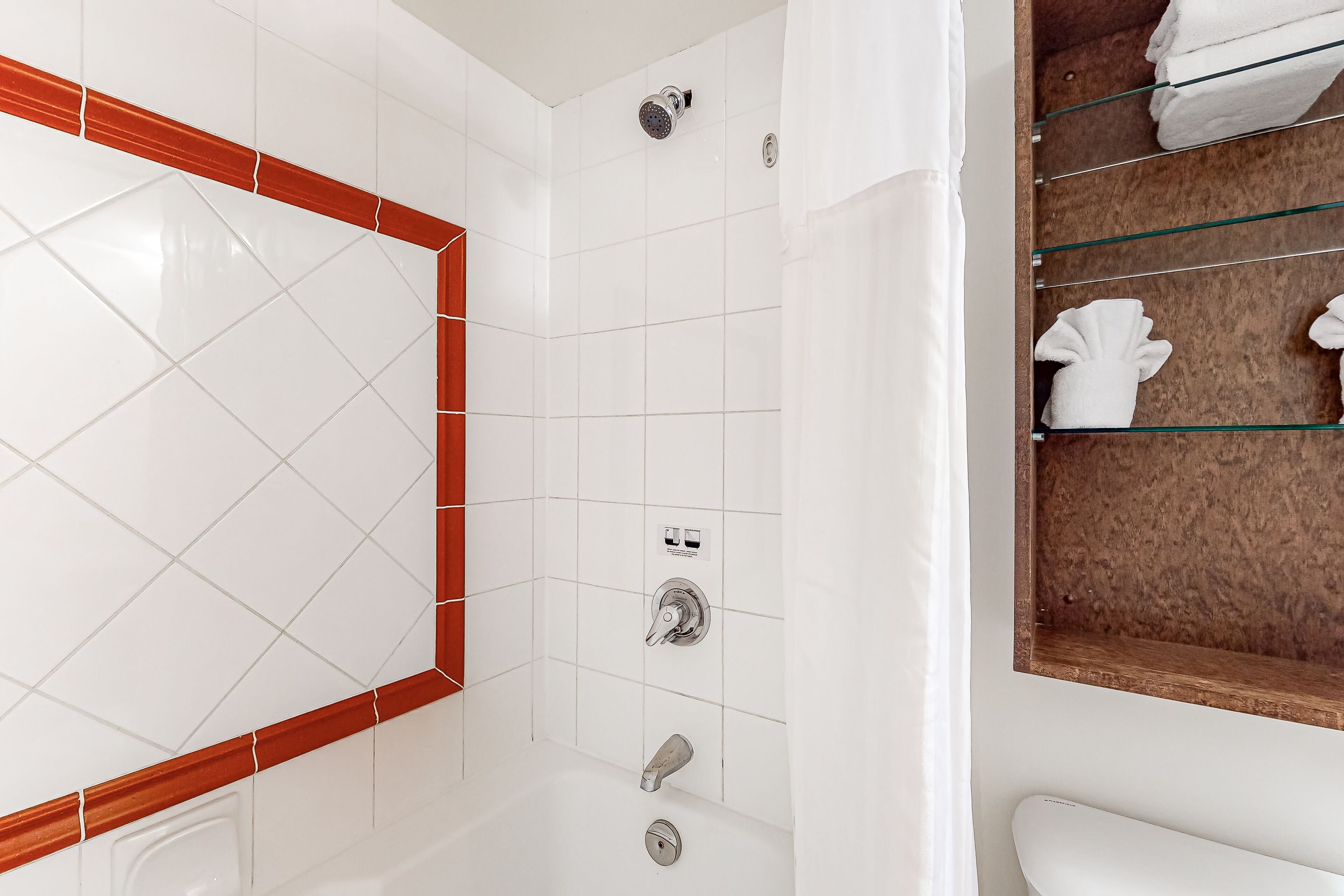 Un baño con paredes de azulejos blancos, un detalle decorativo de madera, una mampara de vidrio en la ducha y un perchero con toallas dobladas.