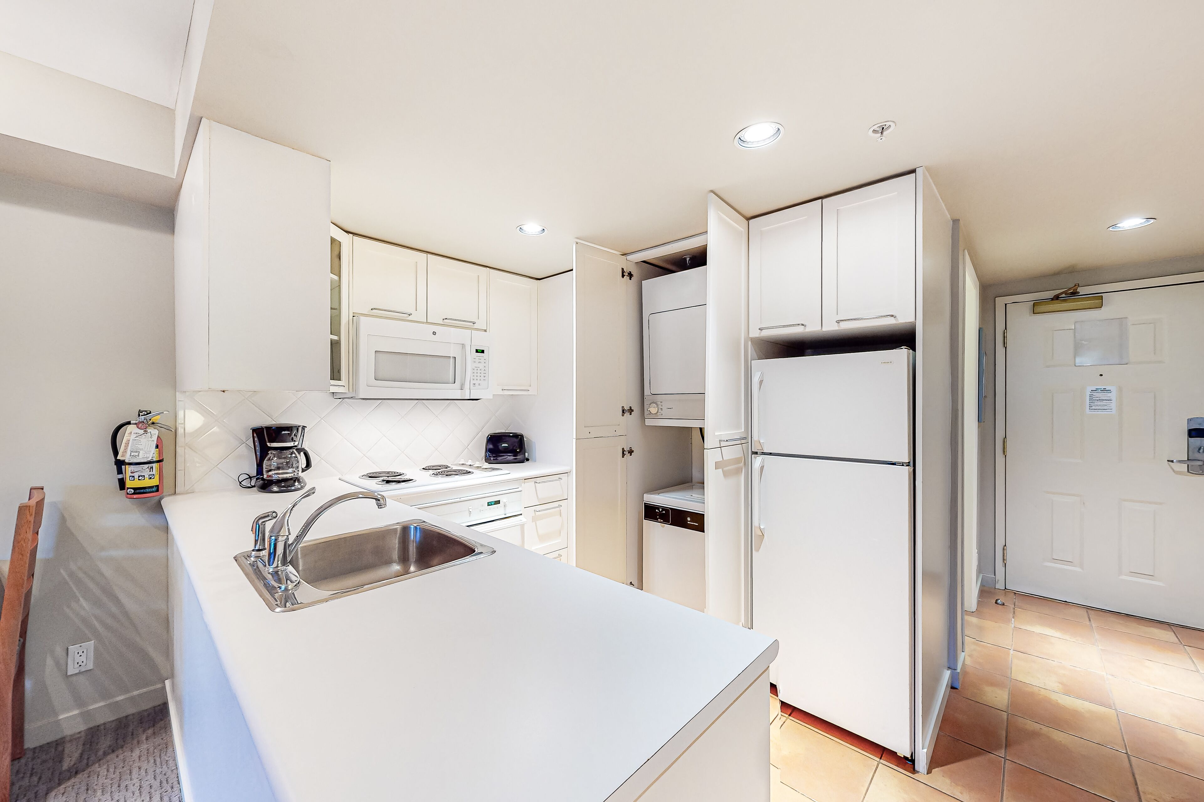 Una cocina moderna con armarios blancos, un lavamanos de acero inoxidable, un microondas, una cafetera y un refrigerador.