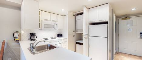Una cocina moderna con armarios blancos, un lavamanos de acero inoxidable, un microondas, una cafetera y un refrigerador.