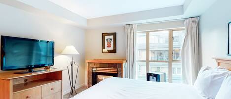A hotel room with a bed, a TV, a fireplace, a window with curtains, and a view of buildings.