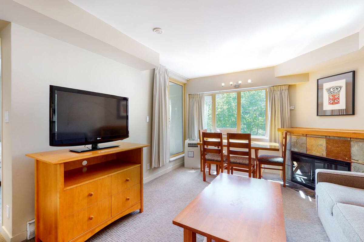 A living room with a TV, wooden furniture, a fireplace, and a large window with curtains.