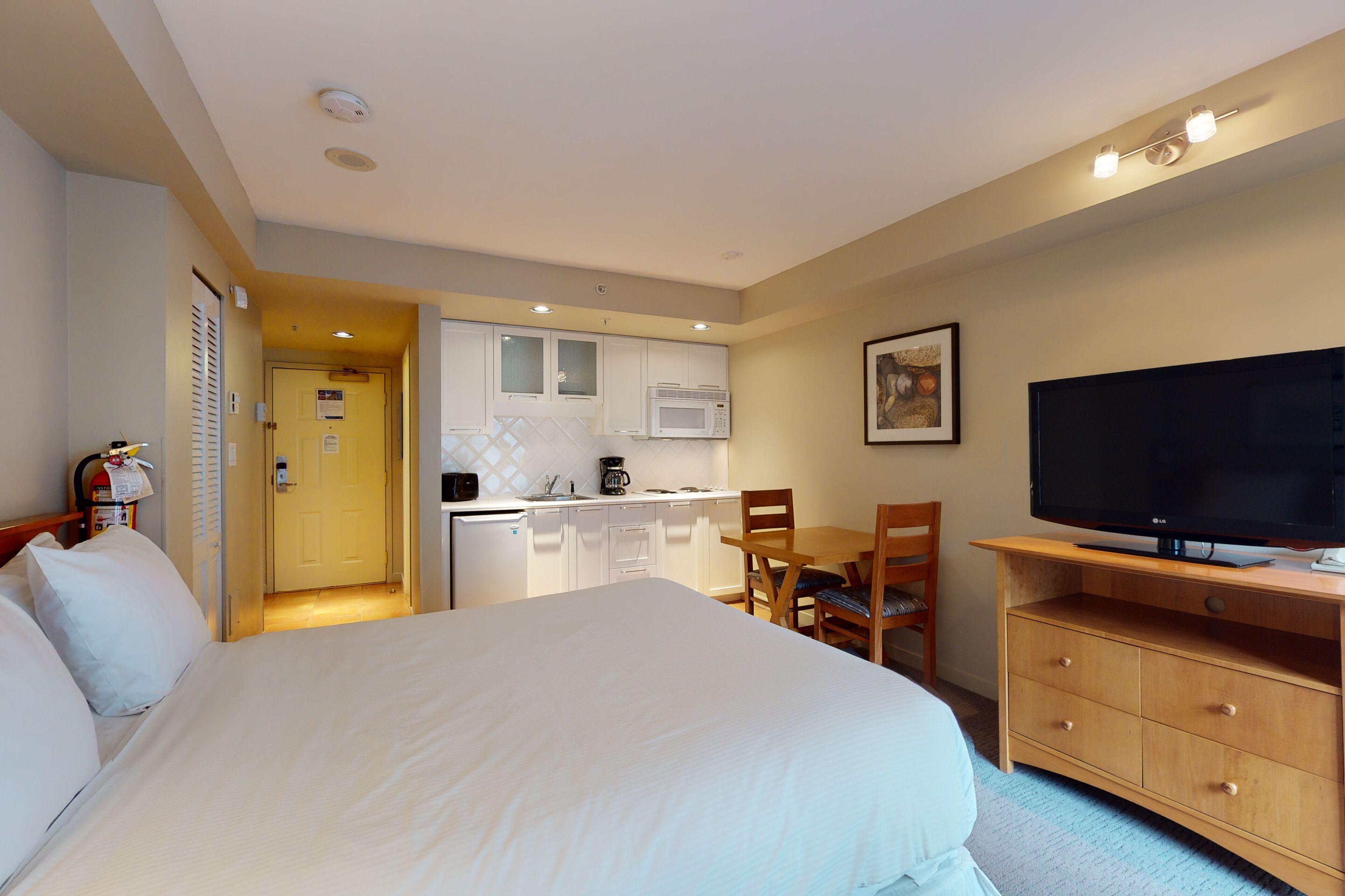 A hotel room with a bed, a TV, a desk, a chair, a small kitchenette, and a door leading to another room.