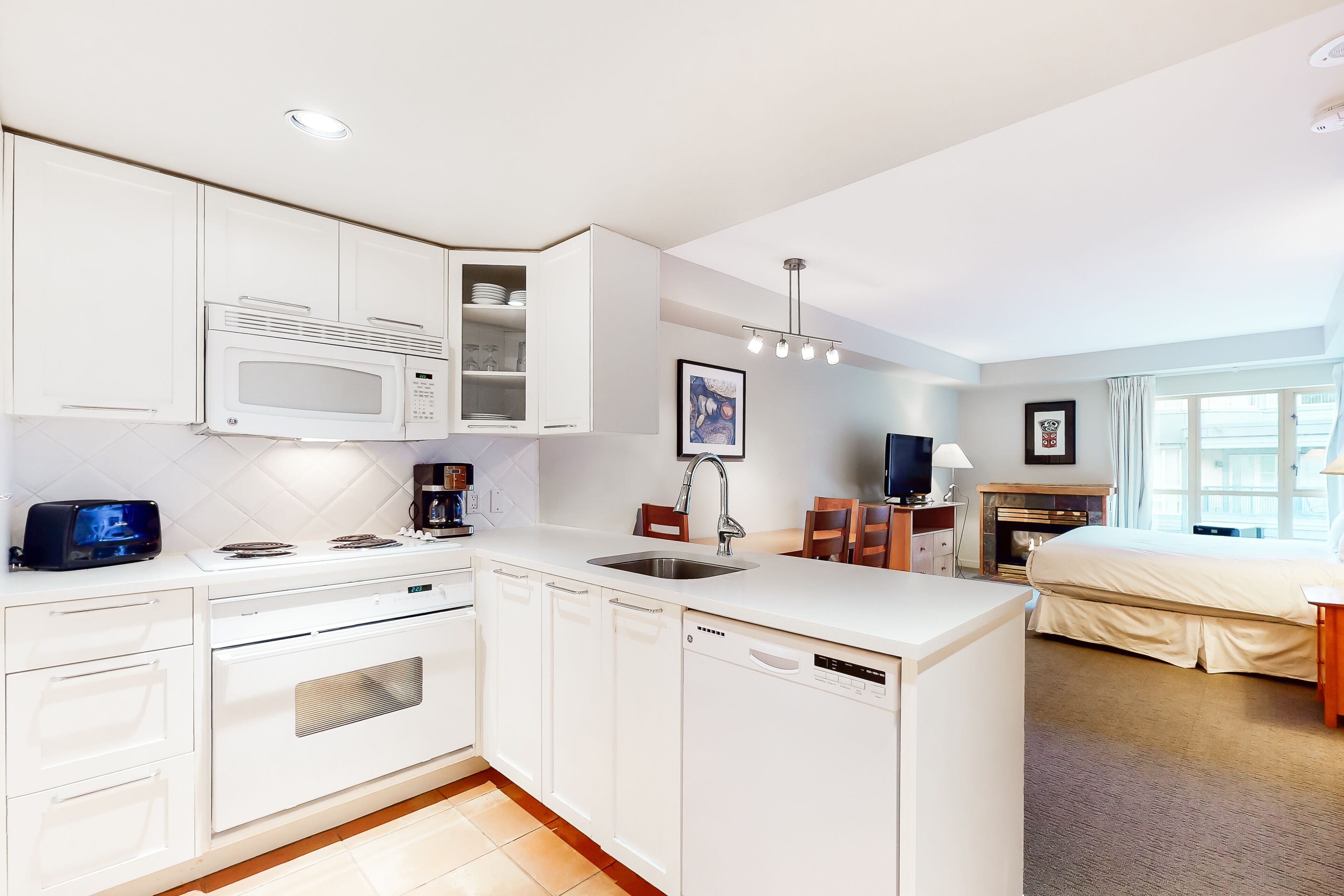 Une cuisine moderne avec des meubles blancs, un four à micro-ondes, une cuisinière et un lave-vaisselle. Il y a également un coin repas avec une table et des chaises, ainsi qu’une chambre avec un lit et une fenêtre.