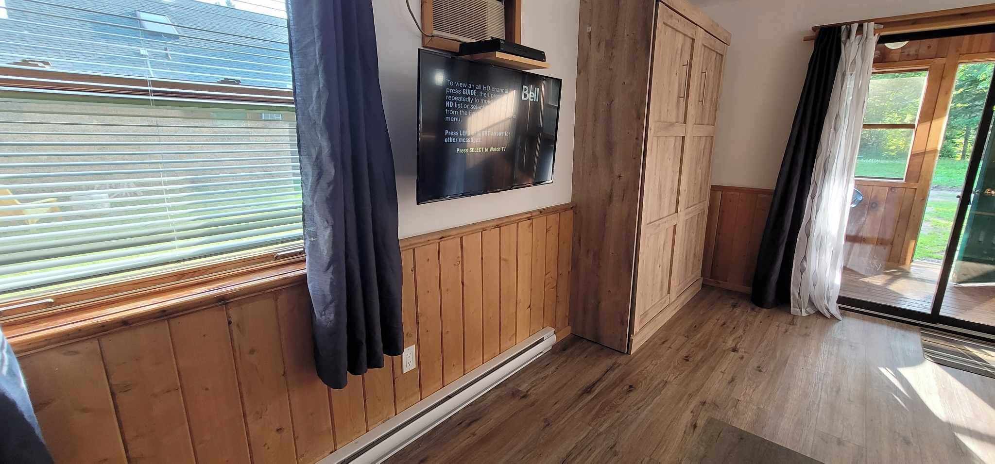 A room with wooden paneling, a mounted TV displaying a guide, a sliding glass door, and a view of greenery outside.