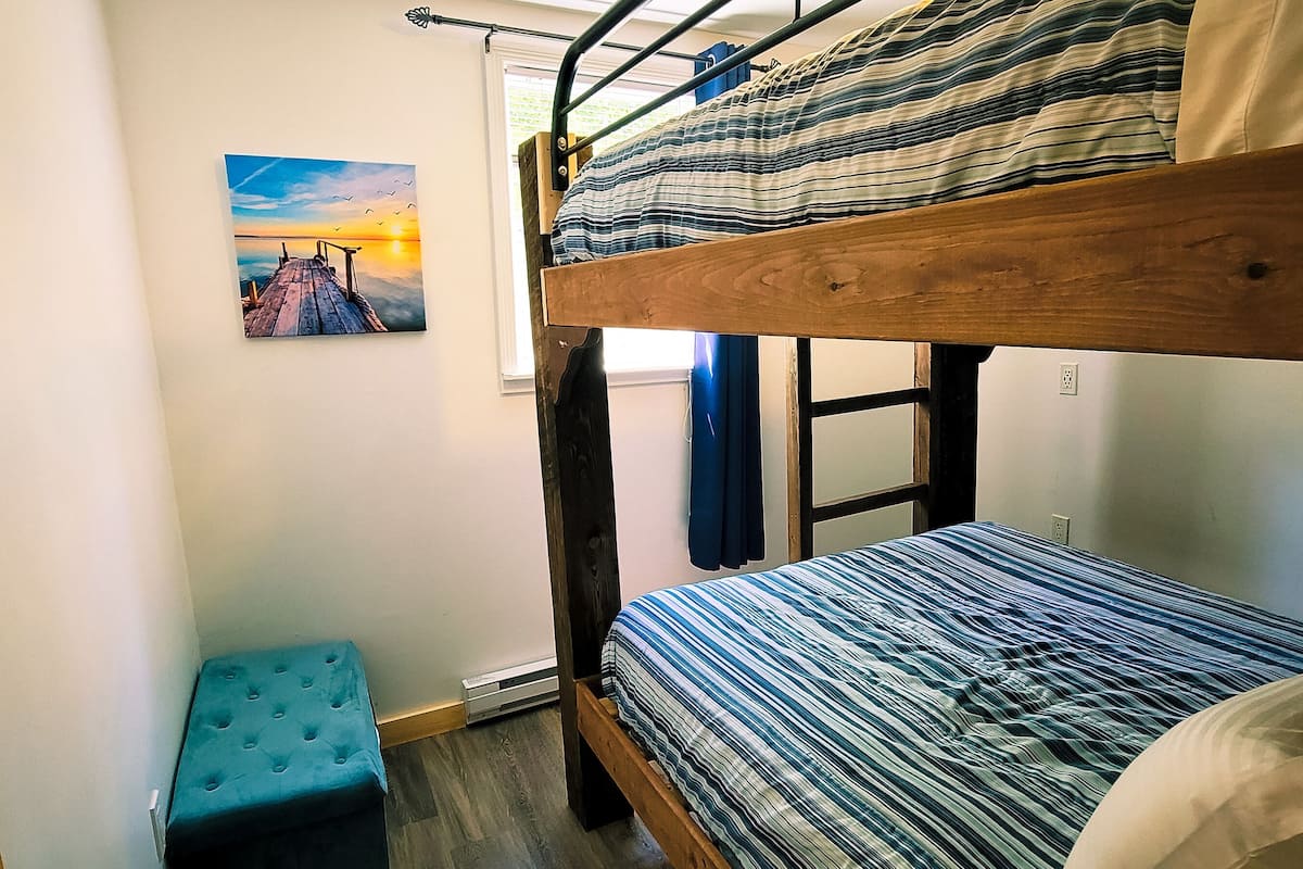 A bunk bed room with a small blue ottoman and a sunset painting on the wall.