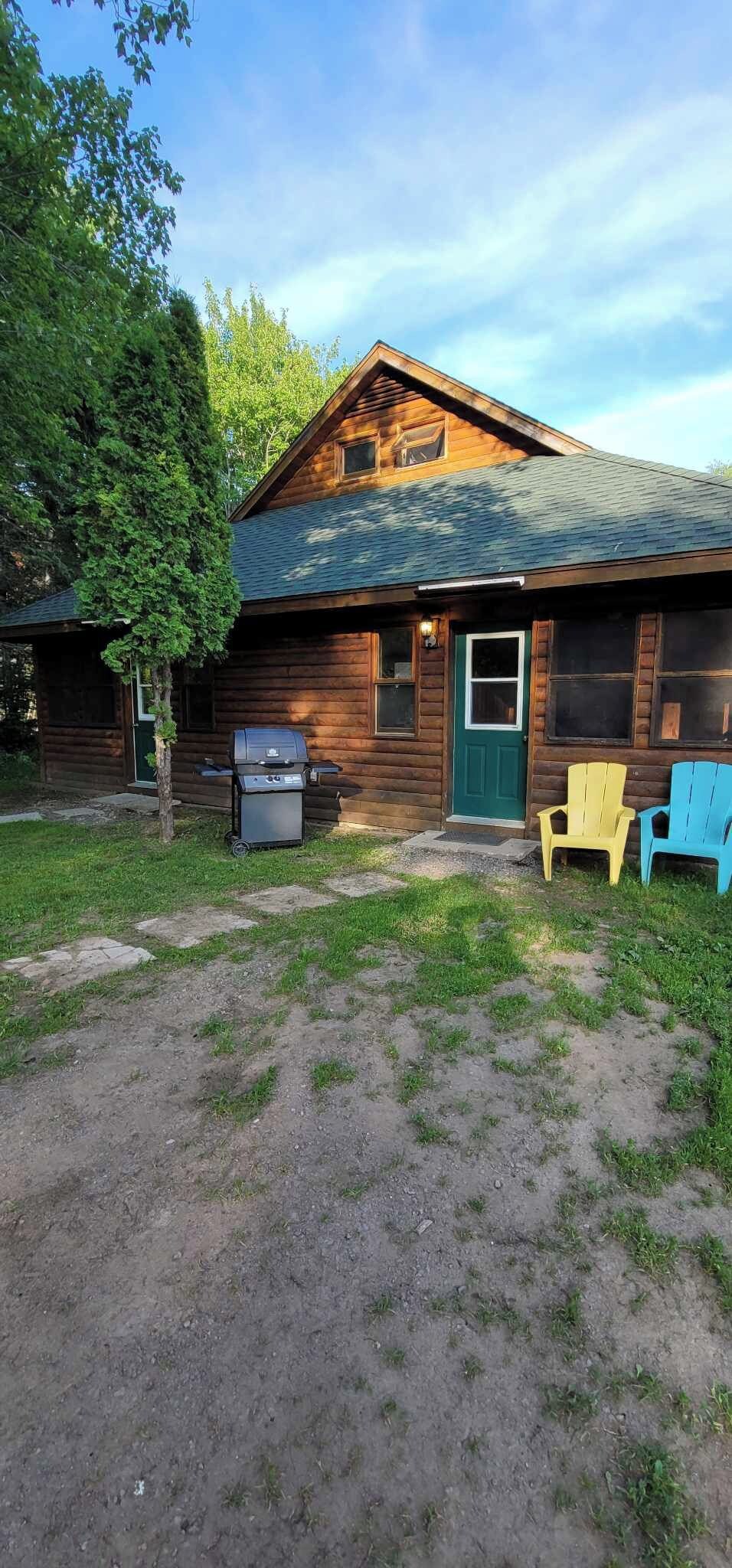 A wooden cabin with a green door, a barbecue grill, and two colorful Adirondack chairs.