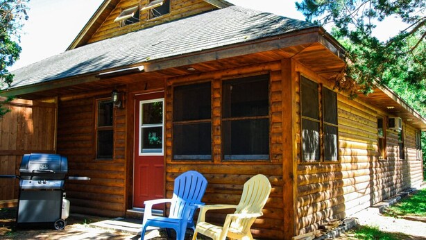 A wooden cabin with a front porch, a grill, and two Adirondack chairs. - Kouch Chalets (Saint Louis de Kent)