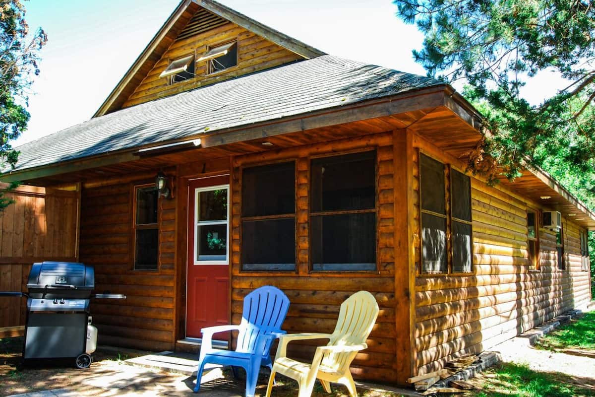 A wooden cabin with a front porch, a grill, and two Adirondack chairs.