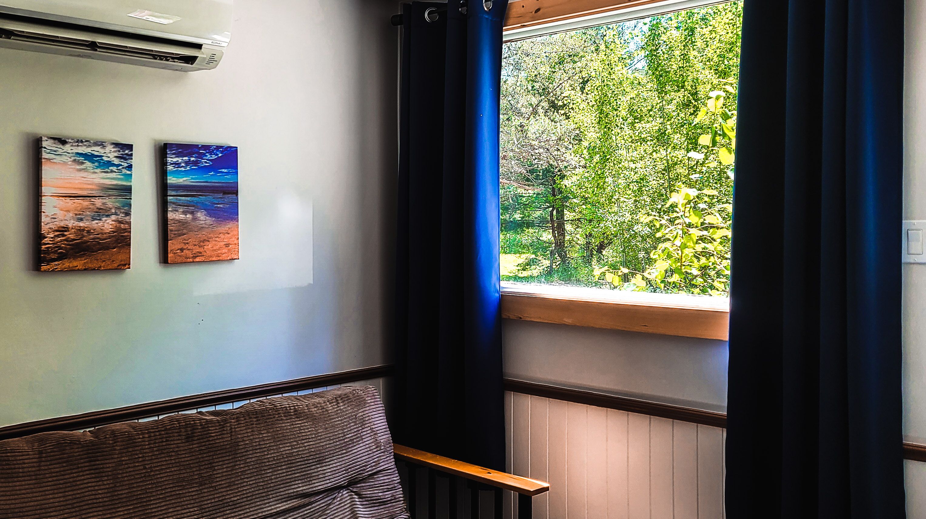 Una habitación con ventana que da a un bosque, un banco y dos cuadros en la pared.