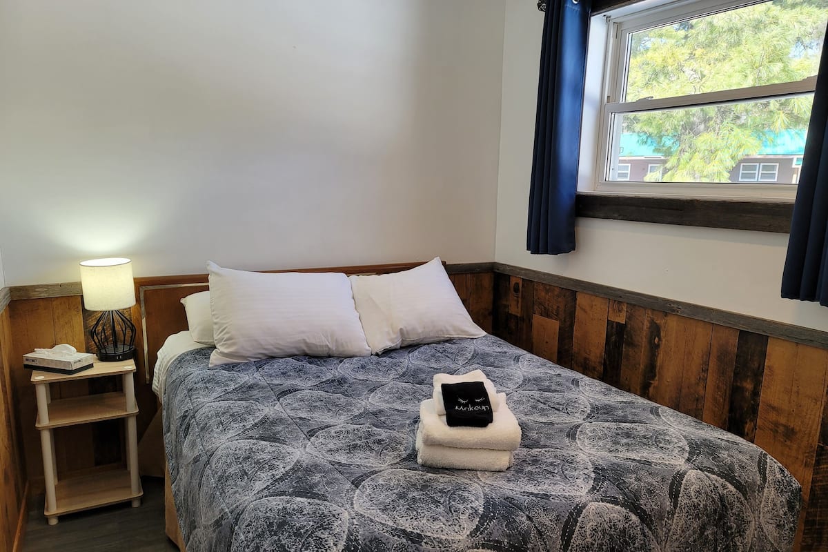 A neatly made bed with a patterned bedspread, two pillows, a bedside lamp, and a small shelf with a tissue box.