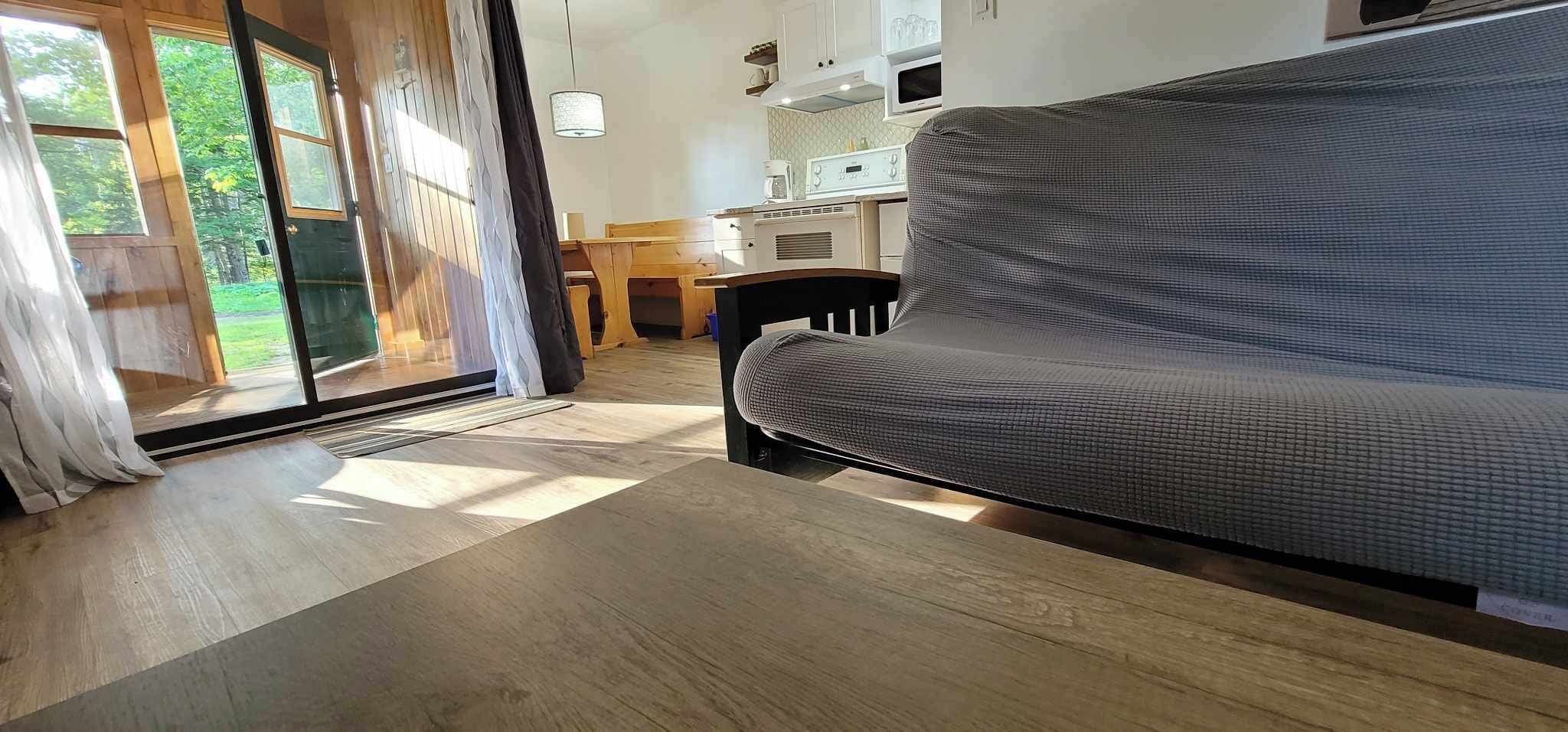 Una sala moderna con un sofá gris, suelo de madera, una zona de cocina con microondas y un ventanal corredero que da a un espacio exterior verde.