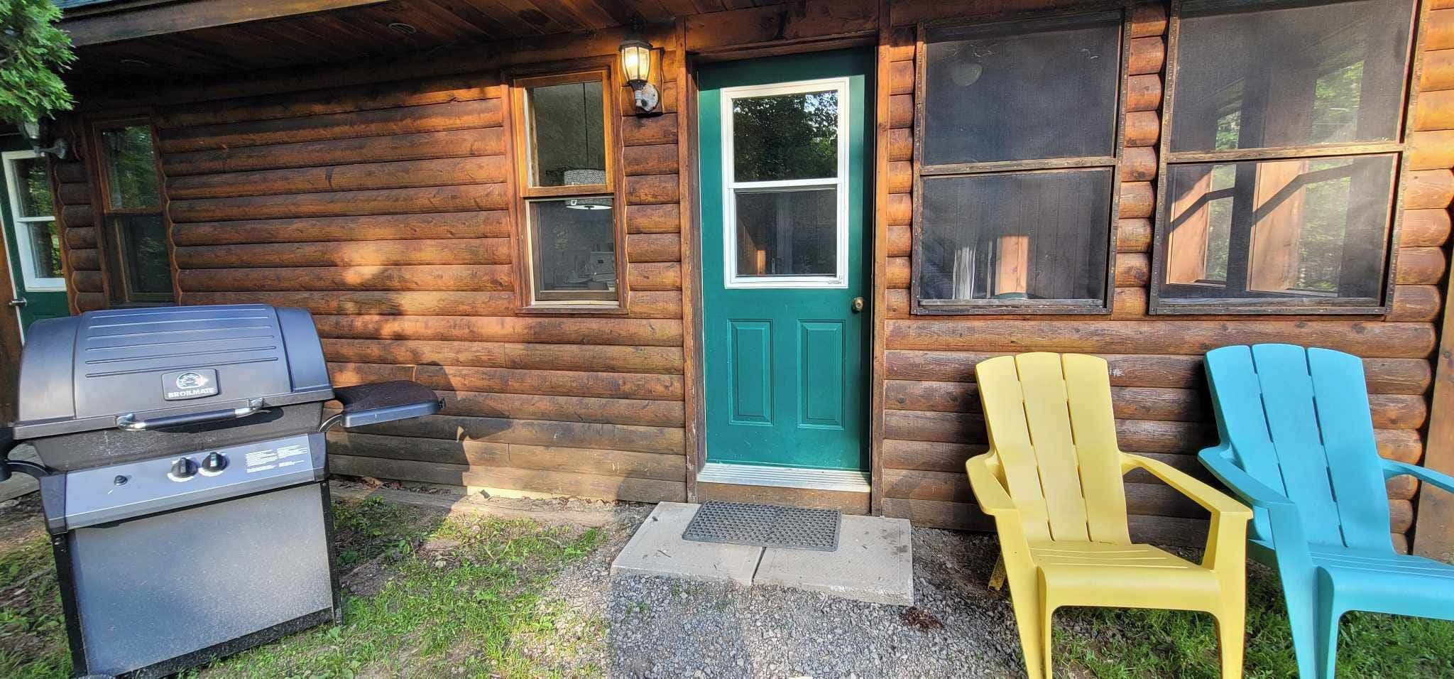 A log cabin with a grill outside, a green door, and two Adirondack chairs.