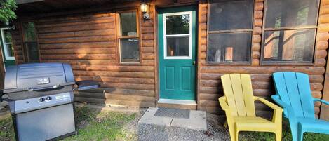 A log cabin with a grill outside, a green door, and two Adirondack chairs.