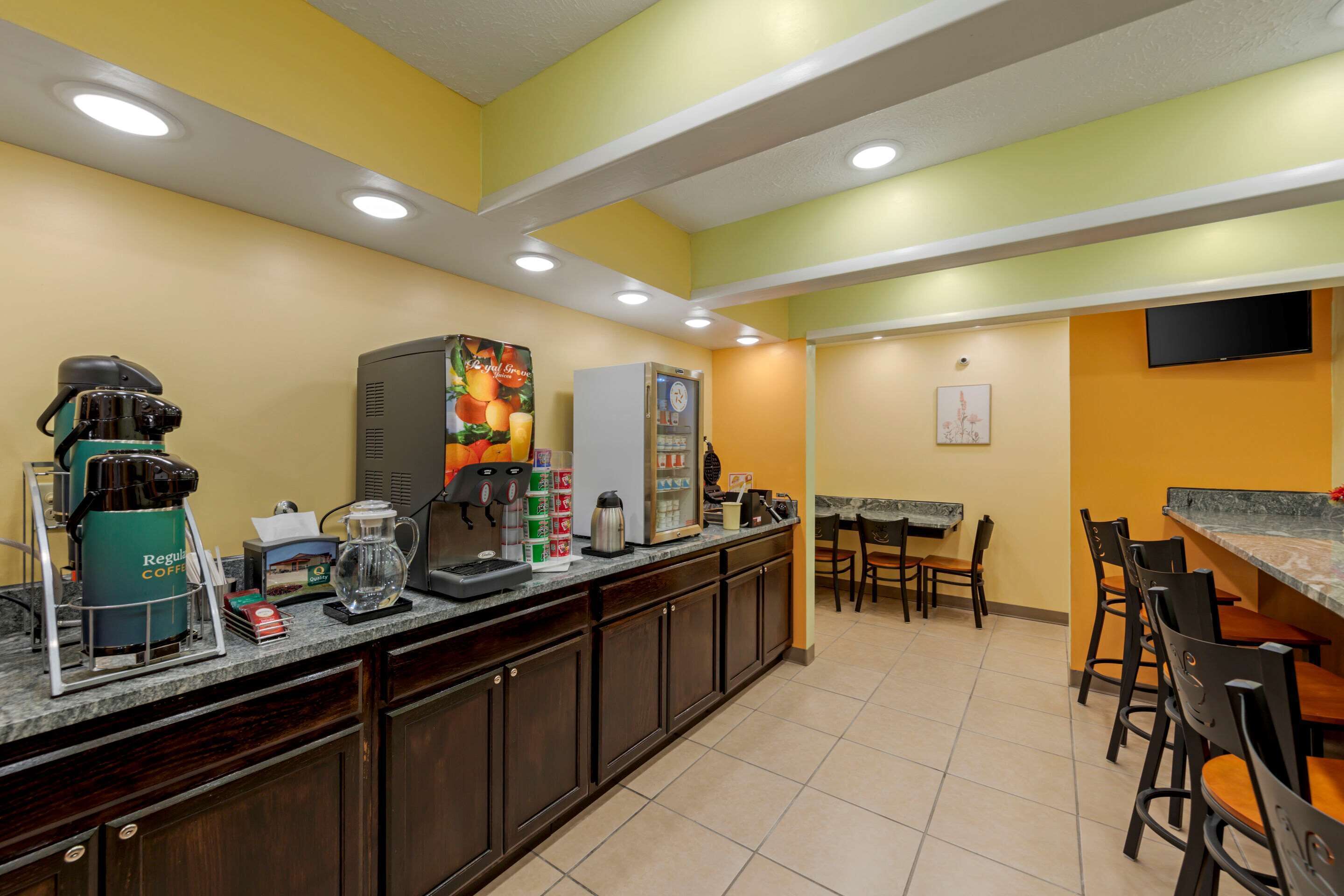A breakfast bar with coffee machines, a refrigerator, and a microwave. There are chairs and a table in the background.