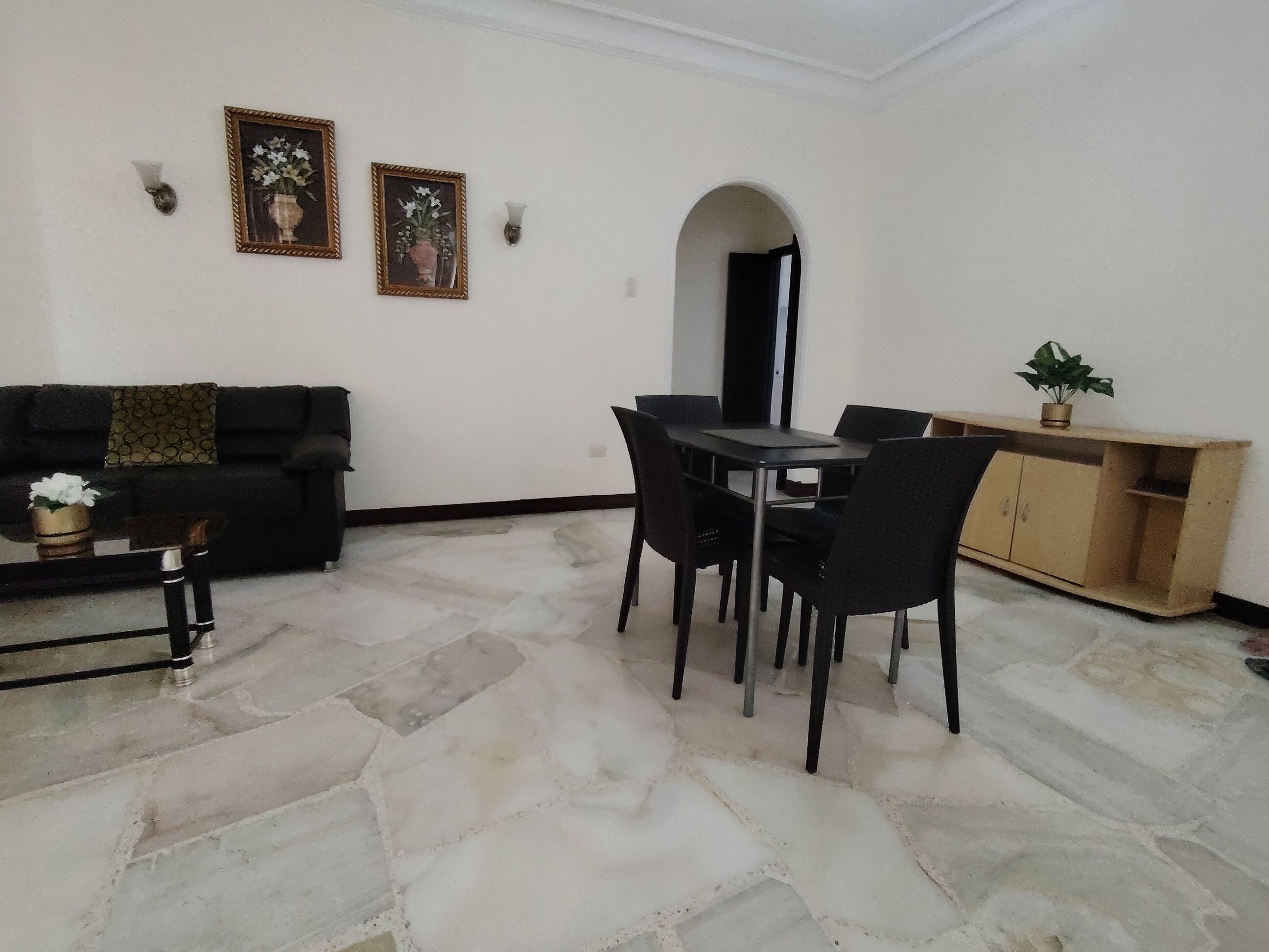 Una sala de estar con un sofá negro, una mesa de comedor con sillas, un aparador de madera y dos marcos de cuadros colgados en la pared.