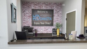 Un mostrador de recepción con un cartel que dice 'Welcome to HD Hotel Dakota Enjoy Your Stay!' y un fondo de pared de ladrillo.