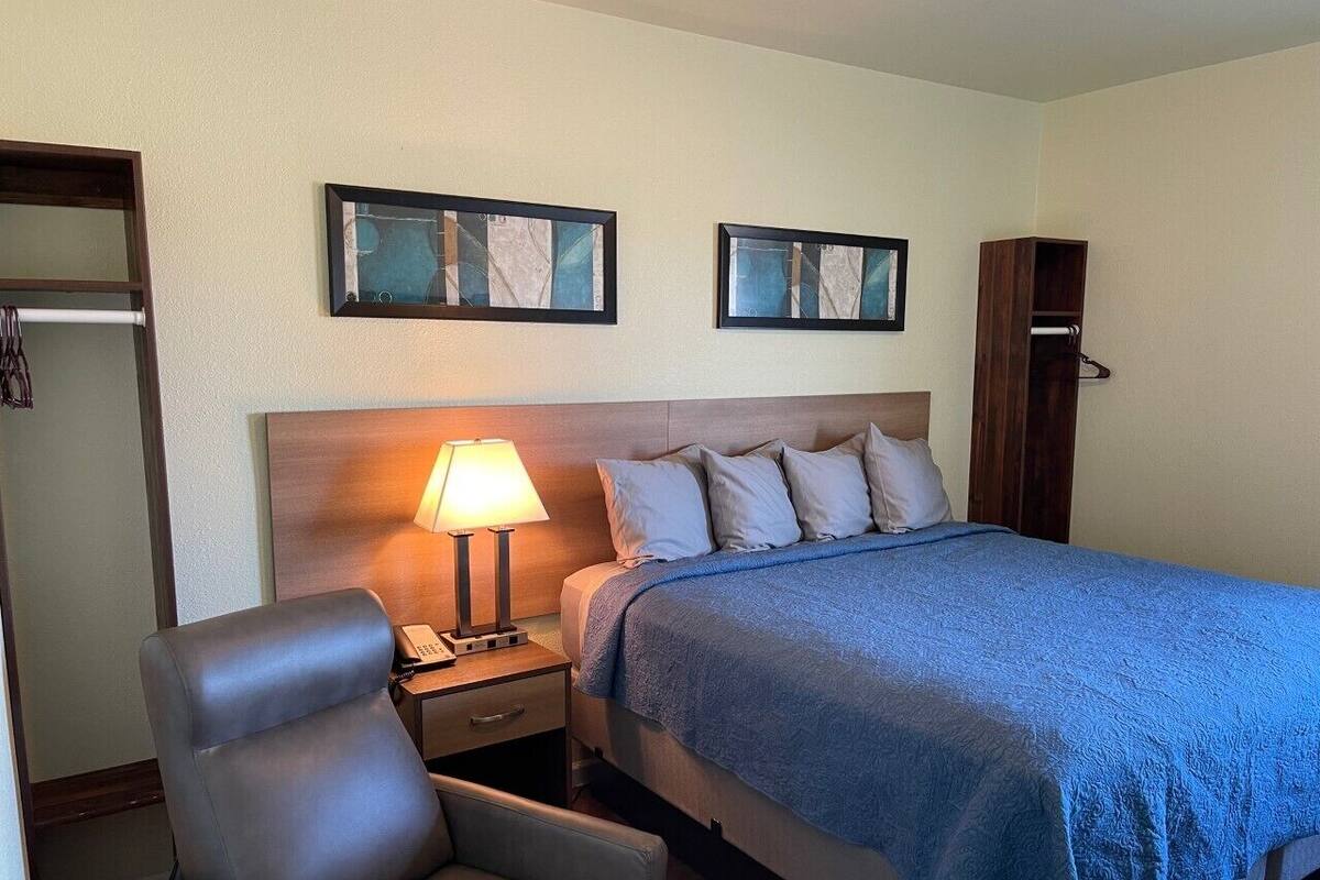 Quarto de hotel com cama, uma cadeira, mesa de cabeceira, abajur e duas imagens emolduradas na parede.
