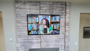 Una sala de estar con un televisor de pantalla plana montado en una pared con textura, dos sillones de cuero, una chimenea y una estantería llena de libros.